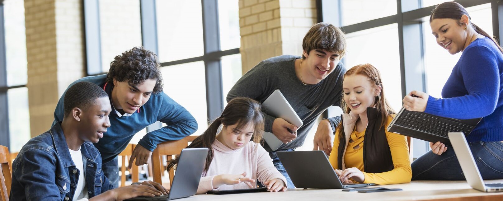 Young adults in a variety of brightly coloured clothing smile as the sit around a table working on their laptops.