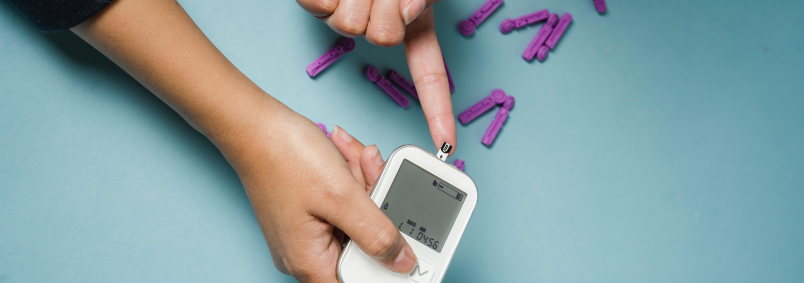 On a turquoise blue background, a hand reaches out to have its finger pricked by a simple white blood monitor held in another hand. Purple plastic clips are scattered across the background.