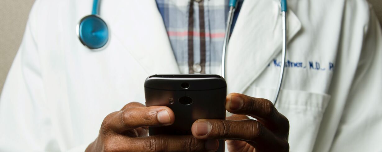 A close up of a pair of hands holding a mobile phone, in the background we can see this person is wearing a white doctor's coat with a stethoscope around their neck.