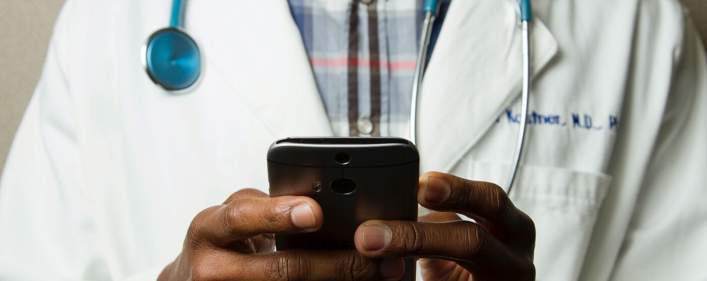 A close up of a pair of hands holding a mobile phone, in the background we can see this person is wearing a white doctor's coat with a stethoscope around their neck.